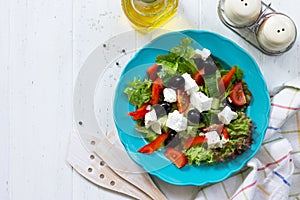 Greek salad with fresh vegetables, feta cheese and black olives in a blue plate on white wooden table.
