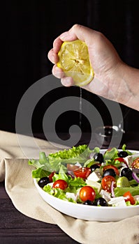 Greek salad in a plate on a dark background. Healthy vegetarian food. The hand squeezes out the lemon juice. Feta cheese, lettuce