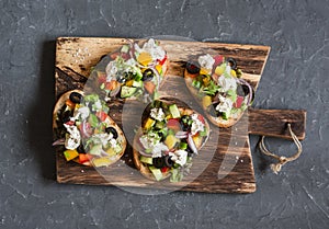 Greek salad style bruschetta on a wooden cutting board, on dark background, top view.