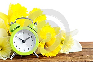 Green alarm clock and spring flowers on table against white background. Time change