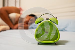 Green alarm clock standing on bed with out of focus man sleeping in bed background. Healthy, sleeping disorder or insomnia concept