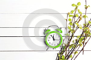 Green alarm clock and young birch branches with first leaves and buds on a white wooden background