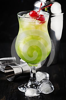 Green alcoholic ocktail with maraschino cherries in hurricane glass, dark bar counter background, selective focus