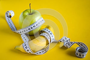 Green apple and banana with white tape measure on yellow background. Healthy lifestyle