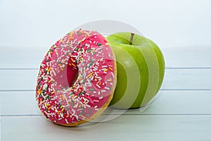 Green apple and donut with strawberry icing and multicolored sprinkles on light wooden table