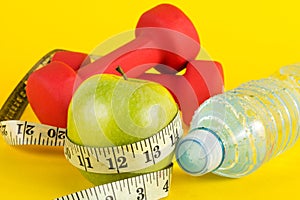 Green apple with measuring tape, red dumbbell and fresh water bottle with drops on yellow background