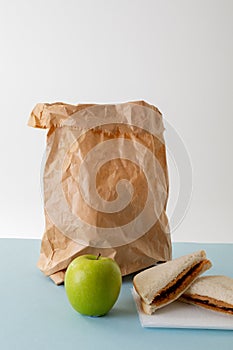 Green apple with paper bag and peanut butter and jelly sandwich on table against gray background