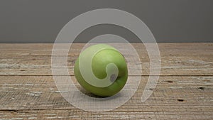 A green apple rolls on a wooden surface on a gray background.