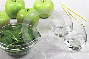 Green apples, a bowl of fresh spinach leaves, two empty glasses and straws