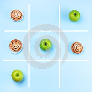 Green apples vs cinnamon buns in tic tac toe or noughts and crosses game, healthy vs unhealthy food concept, top view, square