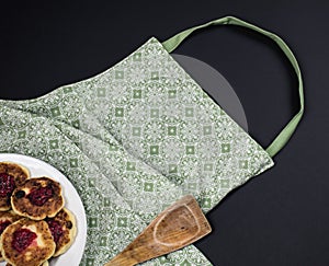 Green apron, wooden spatula and plate with pancakes on a dark background