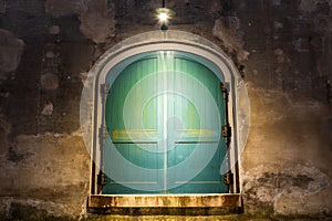 The green arched doors of a warehouse at night with a lone light