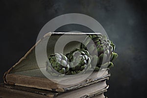 Green artichokes in close-up. Old books. Dark background. Vertical photo. Copy space.