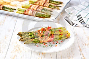 Traditional appetizer, baked bundles of green asparagus wrapped in bacon in a ceramic plate on a white wooden table, close up