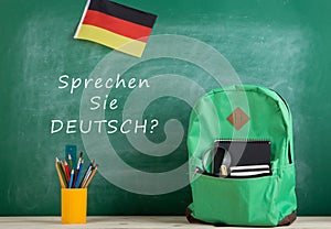 green backpack, blackboard with text "Sprechen Sie Deutsch?", flag of the Germany, school supplies and notebooks