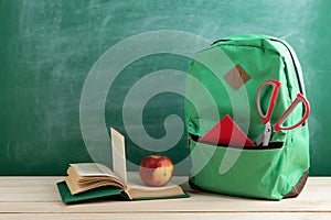 green backpack, open book, red school supplies and apple on the background of the blackboard