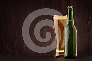 Green beer bottle and glass weizen with golden lager on dark brown wood board, copy space, mock up.