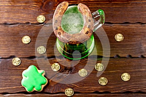 Green beer in a glass mug with gingerbread clover, horseshoe and gold coins on a rustic wooden surface. Festive background for St