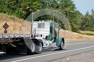 Green big rig day cab semi truck with empty flat bed semi trailer driving to point of loads