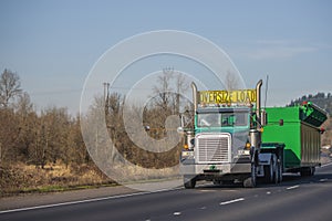 Green big rig semi truck transporting oversize load on step down semi trailer driving on the turning road