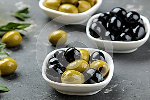 Green and black olives in a white ceramic bowl with leaves on a dark graphite background. close-up