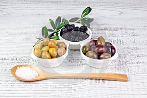 Green, black, pink olives in bowls and a wooden spoon.