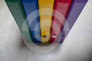 A green, blue, yellow, red and purple binder standing side by side on a grey floor. The concept of work, accounting