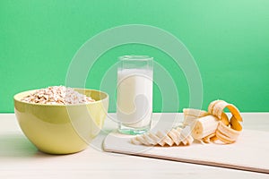 green bowl with dry oatmeal, staak of milk and sliced banana on wooden table