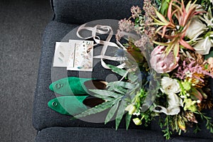 Green bridal shoes with wedding rings on them in focus, green wedding bouquet with pink ribbons and a wedding complimentary lying