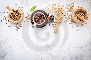 Green and brown decaf unroasted and dark roasted coffee beans in hemp sack bags with scoops setup on white concrete background