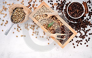 Green and brown decaf unroasted and dark roasted coffee beans in wooden box with scoops setup on white concrete background