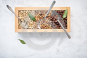 Green and brown decaf unroasted and dark roasted coffee beans in wooden box with scoops setup on white concrete background. with