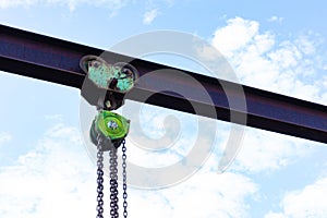 Green chain fall hoist on an I-beam silhouetted against a pale blue sky with clouds