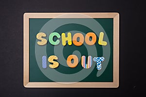 Green chalkboard with wooden frame, text school is out in colorful letters, black wall background