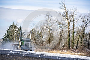 Green classic big rig semi truck with vertical pipes transporting empty flat bed semi trailer running on winter snowy road