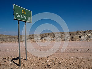 Green color directional road sign pole along unpaved dirt road C14 to Solitaire among rock desert landscape with blue sky