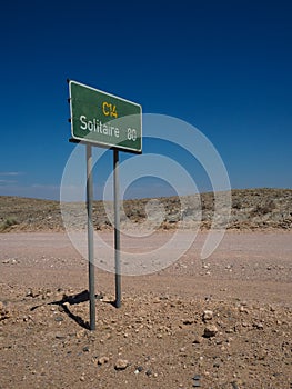 Green color directional road signage pole along unpaved dirt road C14 to Solitaire among rock desert landscape with blue sky