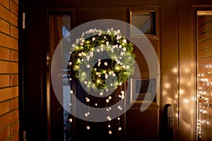 Green color spruce tree tips Christmas Wreath with warm lights and white felt pom-poms on home entryway.