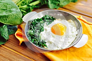 Green cooked spinach with fried egg on iron pan plate. Fried eggs healthy breakfast on wooden table background on orange napkin