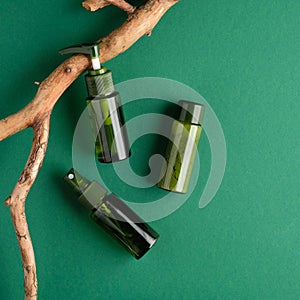 Green cosmetics bottles and wooden branch on green background. SPA natural organic beauty products set. Flat lay, top view