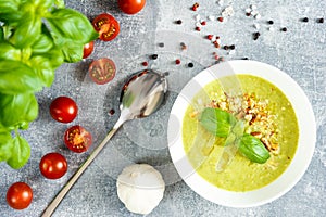 Green cream soup on the white bolw, spoon, garlic, cherry tomatoes, black and red grainy pepper, grey background