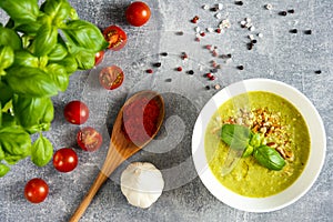 Green cream soup on the white bowl, basil, cherry tomatoes, wooden spoon with red pepper, garlic