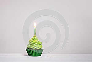 Green cupcake with sprinkles and lit birthday candle on a white grey background.