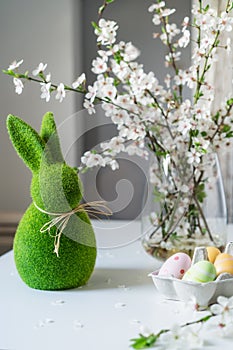 Green easter bunny with blooming flowers on tree branch in the vase and colored easter eggs in container on the white