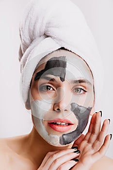 Green-eyed girl is doing spa facial procedure on white background. Portrait of woman in towel with clay mask.