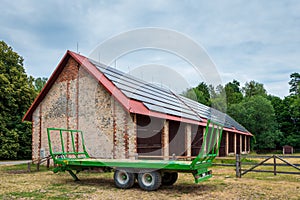 Green farm trailer parked in front of a large barn. Solar panels installed on the roof of the barn