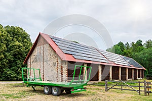 Green farm trailer parked in front of a large barn. Solar panels installed on the roof of the barn