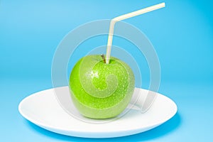 Green fresh apple with water drops. Juicy apple with straw on the plate on cheerful blue background. Natural apple juice concept