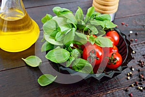 Green fresh leaves of organic basil and small ripe tomatoes, oil and pepper on a wooden background