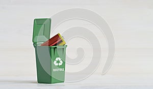 Green garbage sorting container, with an open lid, batteries, and an ecology icon. Close-up, white wooden isolated background. The